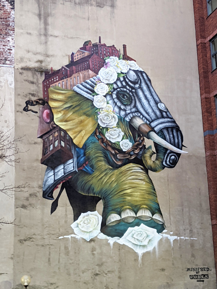 Mural of an armoured elephant with an industrial city on its back and a headdress made of white roses