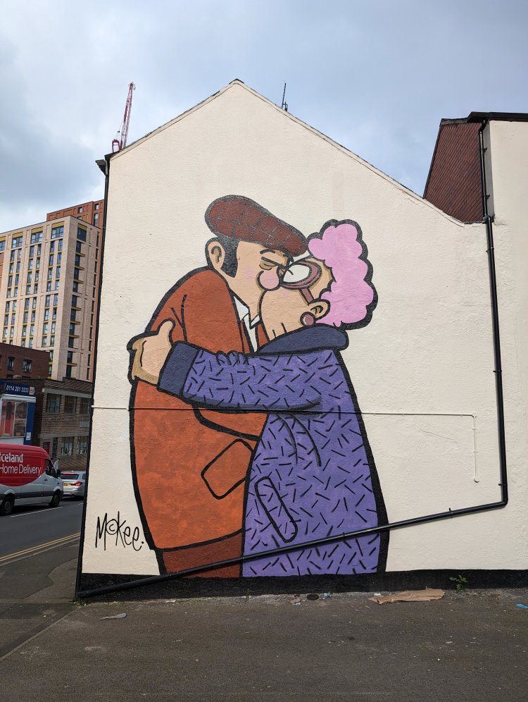 Large mural of an elderly couple embracing in a kiss; the man is wearing a jacket and flatcap, the woman is wearing a wollen coat and spectacles