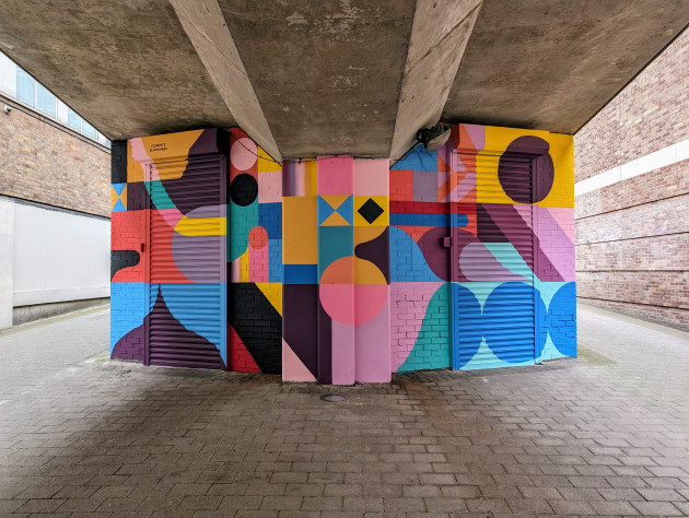 Colourful abstract mural of different shapes on a wall and shutters below a ramp leading up to a car park