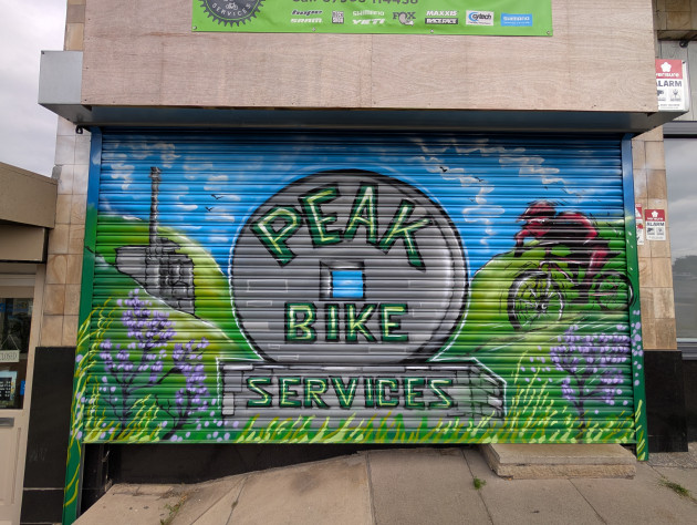 Shop shutters painted with a peak district landscape, a large wheel-shaped millstone takes centre place with the words Peak Bike Services