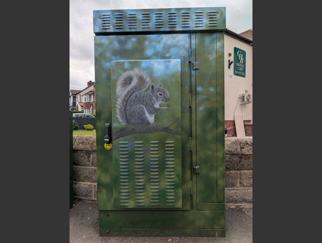 Street cabinet painted with a mural of a grey squirrel perched on a tree branch, it is holding an acorn between its front paws
