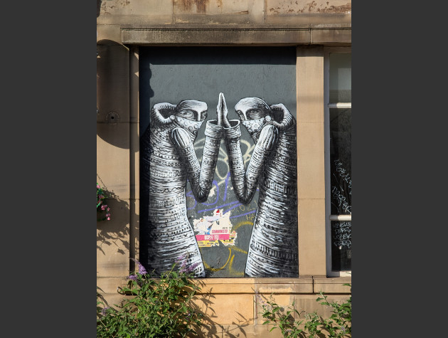 Black and white illustrative mural painted in a boarded up window, featuring two cloaked figures, raised hands touching each other, peering out of the mural