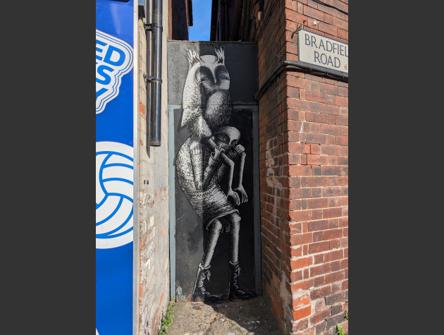 Black and white illustrative mural sandwiched between two buildings, it features an arched back figure giving a piggyback to a long limbed owl