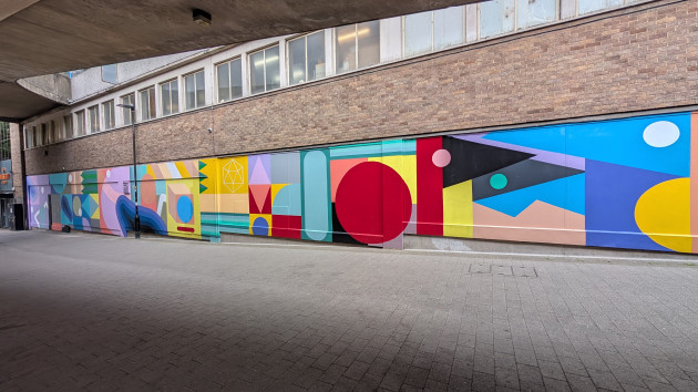 Colourful mural stretching along a wide side wall of a building. The mural consists of various differently coloured shapes.