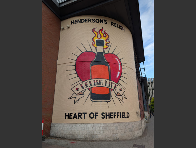 Mural featuring a large bottle of Henderson's Relish, flames rising from its top and a red heart with protruding rays behind, a banner over the bottle reads Relish Life and the words Heart of Sheffield below