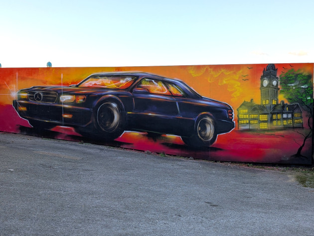 Graffiti mural featuring a classic dark blue Mercedes-Benz coupe set against a fiery orange and red sunset, to the right, a stylized depiction of Sheffield Town Hall