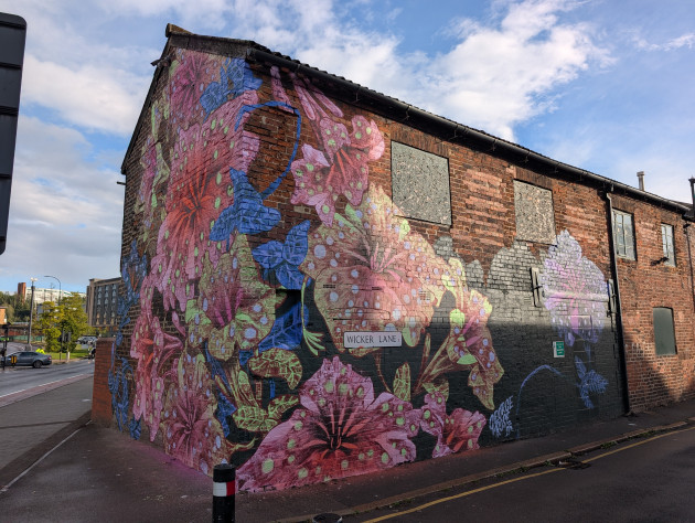 Mural covering the corner of an old red brick building featuring oversized, stylized flowers in vibrant shades of pink, blue and lime green
