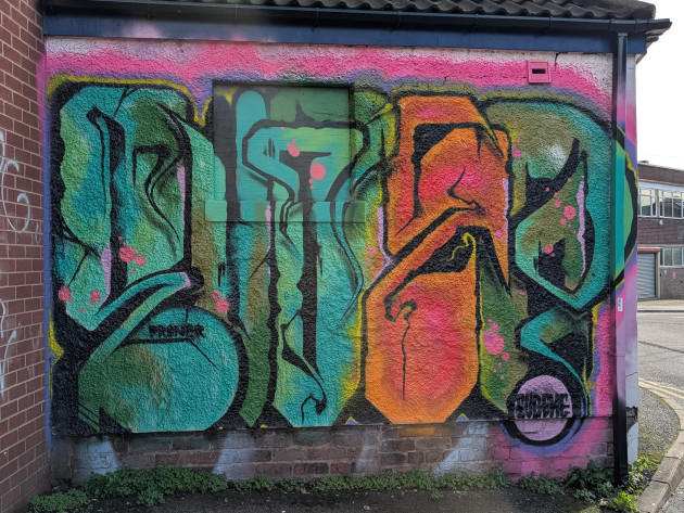 A vibrant graffiti mural on a textured wall featuring large, abstract lettering in teal, green and orange. The letters are defined by thick black outlines and accented with bright pink splatters, all set against a fuzzy pink and black border.