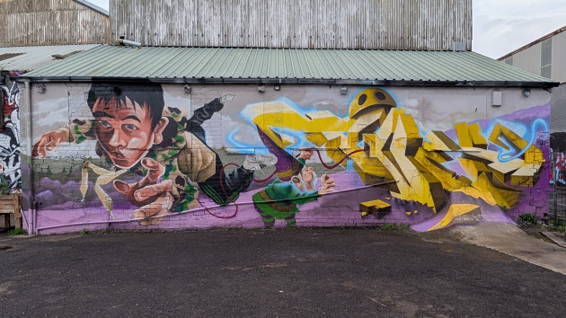 A large surreal mural featuring a track suited boy in free fall facing outwards, while a gnome to the left of the boy appears to be magicing up 3 dimensional wild style lettering which appear to be being eaten by a yellow pacman character