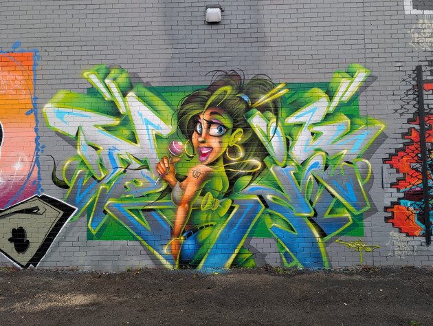 Graffiti mural of a young woman seen from behind with her head turned towards the viewer, she holds a round pink lollipop in her right hand and is surrounded by wild style lettering