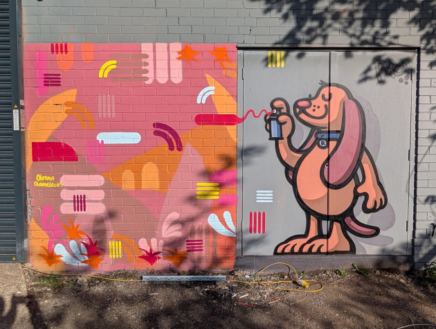 A two-part mural across a brick wall and a metal door. On the right, a cartoonish brown dog with a blue collar and a smile holds a spray can. A red line of "paint" from the can leads to the left side of the mural, which consists of an abstract patchwork of pink and orange geometric shapes.