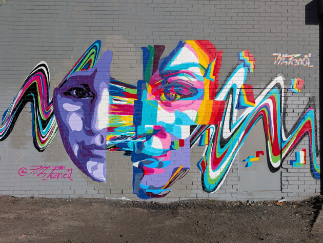 A vibrant graffiti mural featuring a large human face split vertically. The left side is painted in shades of purple and lavender, while the right side is a colourful, fragmented "glitch" of bright red, yellow and blue. A wavy, multi-coloured ribbon flows horizontally behind the face.