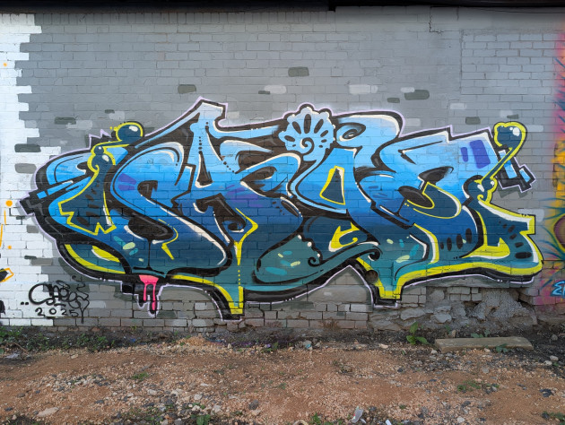 Graffiti piece using wild style writing to spell out Sage with a blue to green fade and yellow highlights, a small section features drips of vibrant pink oozing from the letter S