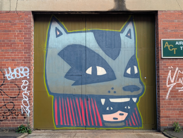 Mural of a masked figure with long, thick red hair, wearing a feline creature's head as a hat. The feline creature has tiger like markings, pointed ears and fanged teeth.