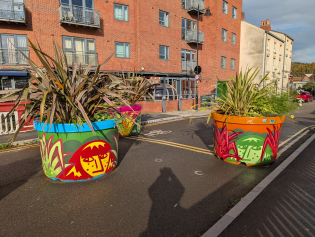 Three large street planters feature bold murals by Ethan Lemon. The artwork shows stylized, freckled faces peeking through vibrant foliage. One planter displays a red-haired figure framed by green leafs; another shows a green face framed by red plants.