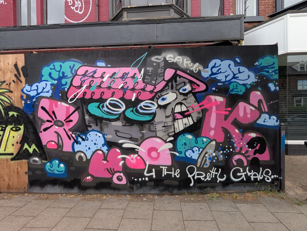 Surreal graffiti mural painted in a cartoony style; it features an anthropomorphic grey brick house, with pink roof and chimnel surrounded by trees, bushes and the lettering R U OK