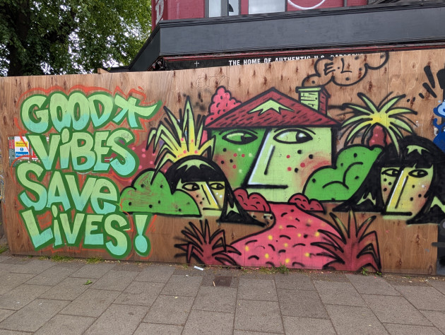 Graffiti mural with the words 'Good vibes save lives' and a surreal picture featuring freckled faces surrounded by plants and a central freckled face in the shape of a house