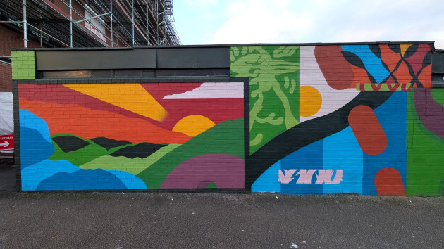 Vibrant mural on a brick wall. The left side features a geometric sunset with a yellow sun over rolling green hills under a red and orange sky. The right side transitions into abstract blocks of blue, green and yellow.