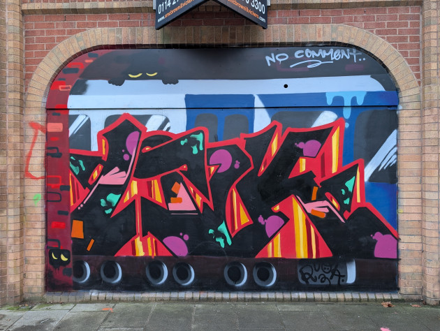 Mural of a train carriage seen passing through a tunnel. The carriage is covered in wild style graffiti. Mysterious faceless eyes can be seen in the shadows of the tunnel.