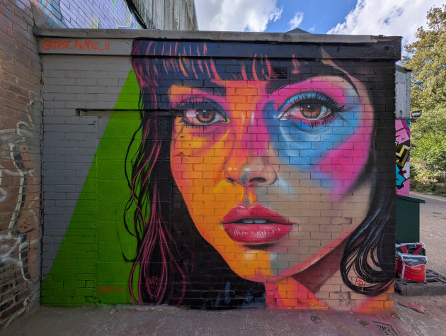 Vibrant mural of a young woman's face. The right side of the face is glowing orange and shades of blue and pink frame her left eye.