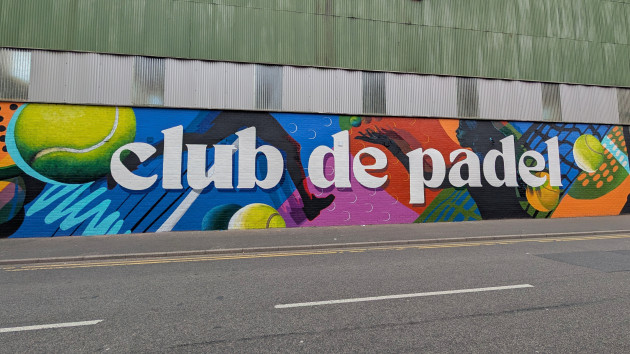 Wide wall mural with the writing "club de padel" written across the middle. In the background are silhouettes of padel players and realistic looking padel balls.