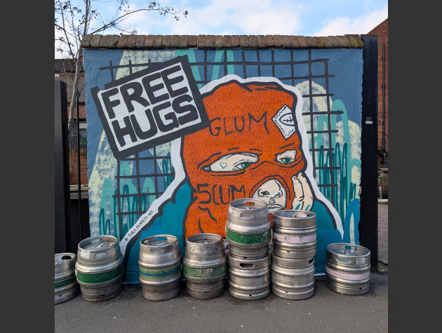 Mural of a tattooed figure in an orange balaclava with 'Glum Scum' stiched into it. A sign states 'Free Hugs'.