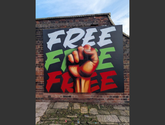 Mural of a raised clenched fist over the word "FREE" repeated three times in white, green, and red on a black background. The art is mounted on an outdoor brick wall.