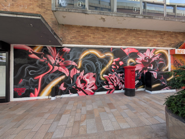 Mural featuring a side profile of a woman's face outlined by pink lighting, surrounded by pink flowers and light drifting smoke. A glowing thread weaves between the other elements of the artwork.