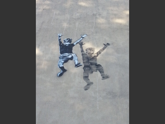 Small stencil artwork of a leaping boy, shadow behind them, high up on the side of a two-storey wall