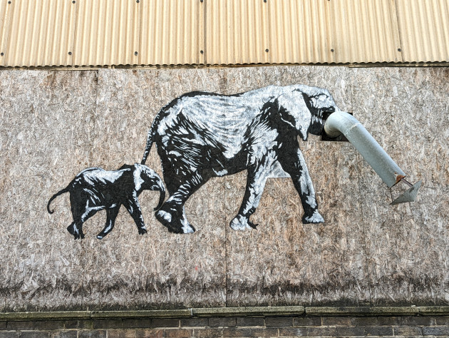 Black and white mural of an elephant with her child in tow, the adult's trunk is a defunct pipe from the building