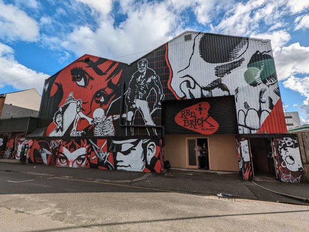Huge red, black and white mural covering the facade of a three-storey high warehouse featuring various characters and symbols