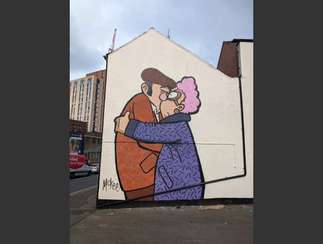 Large mural of an elderly couple embracing in a kiss; the man is wearing a jacket and flatcap, the woman is wearing a wollen coat and spectacles