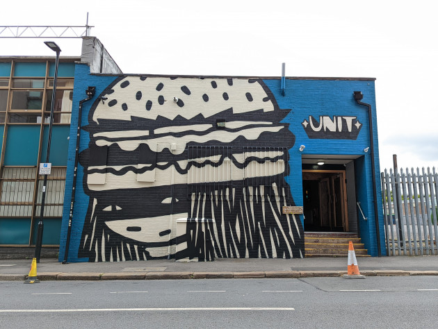 Cartoon style mural of a masked woman wearing a burger for a hat on the facade of the Unit restaurant