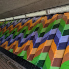 Close up of the mural featuring an orange, green and purple zig-zag pattern, the wall surface is slanted with frequent columns of raised bricks