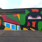Mural section consisting of two parts. The left hand side features an abstract representation of Endcliffe Park café. The right hand side is a close up of an eye part of an abstract face formed of bold black strokes and red, orange and green fill.
