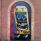 Mural of the front of a train, appearing to emerge from a tunnel, helped framed by the brick work of the wall the mural is painted on. The front of the train has angry eyes and an open mouth spluttering water.